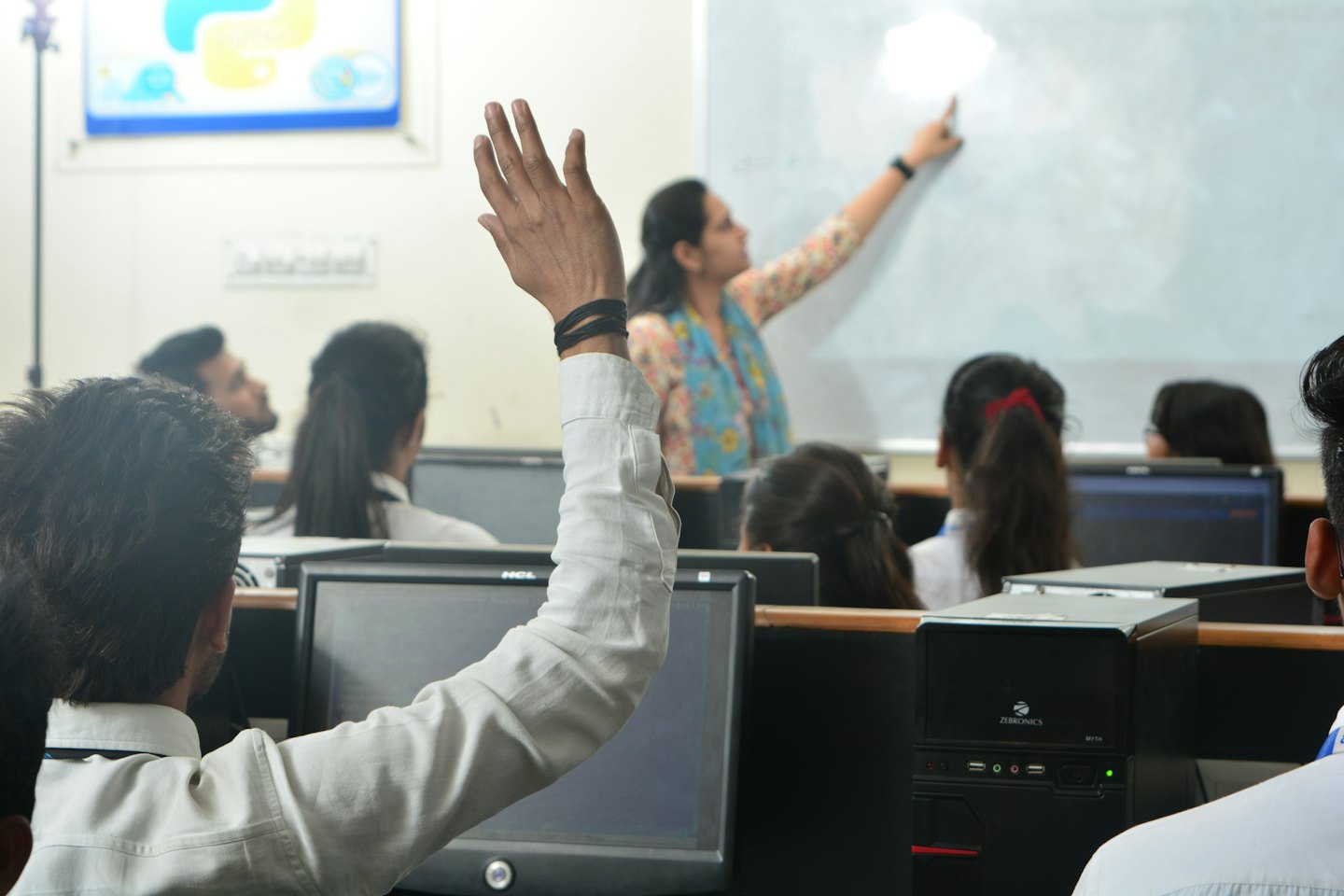 Teacher points at whiteboard in a busy computer lab classroom.